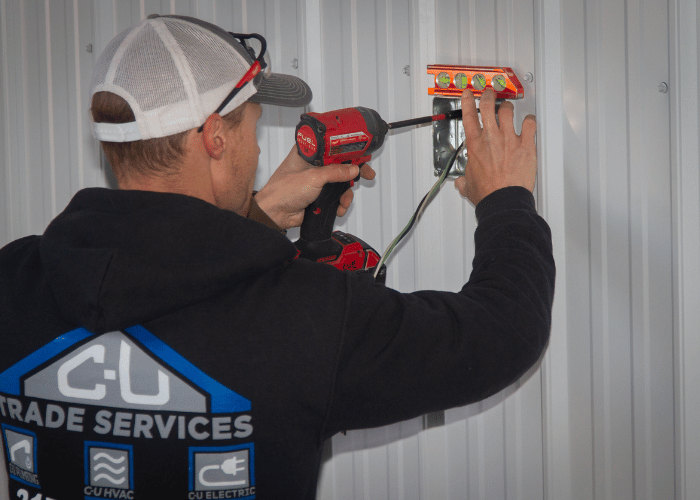 C-U Trade Services electrician using a power drill to install a junction box on a metal wall, with exposed wiring and a level for alignment.