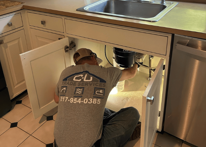 C-U Trade Services technician repairing plumbing under a kitchen sink, working with a garbage disposal unit and illuminated work area inside an open cabinet.