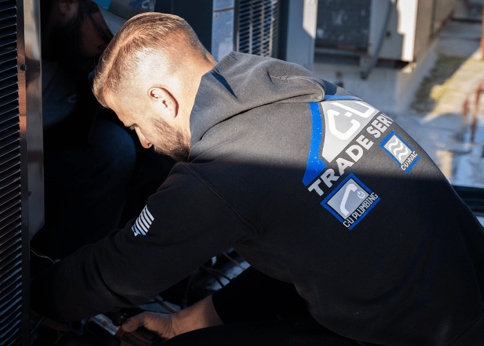 C-U Trade Services technician in branded hoodie working on HVAC equipment outdoors, performing maintenance or repair on a commercial or residential unit.
