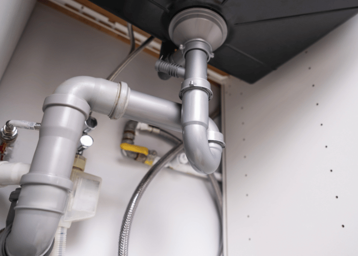 Close-up view of plumbing under a kitchen sink, showing a modern PVC drain trap assembly, water supply lines, and connections to appliances.