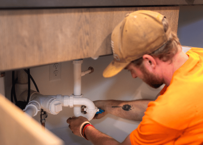 Plumber working under a kitchen sink, tightening a white PVC pipe connection using a wrench, with visible drain assembly and supply lines.