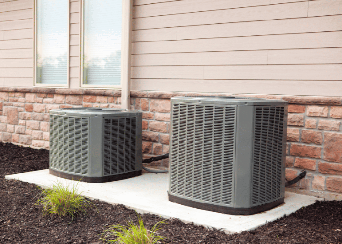 Two outdoor air conditioning condenser units installed on a concrete pad next to a house with brick and siding exterior, part of a central HVAC system.