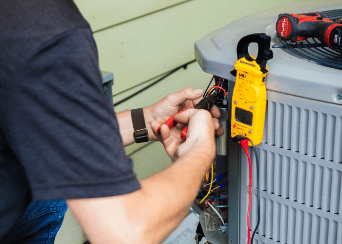 Close-up of an HVAC technician using tools and a multimeter to test and repair the wiring inside an outdoor air conditioning unit during maintenance.