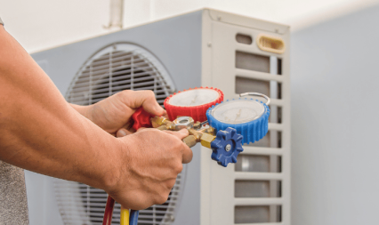 This image features a person's hands adjusting gauges on a brass manifold, used for monitoring pressure in an air conditioning system. The gauges are color-coded, red and blue, each with detailed scales indicating pressure levels. In the background, the air conditioning unit's fan and outer casing are visible, suggesting maintenance work is being performed. The focus is on the hands and gauges, emphasizing the action of tuning or diagnosing the system.