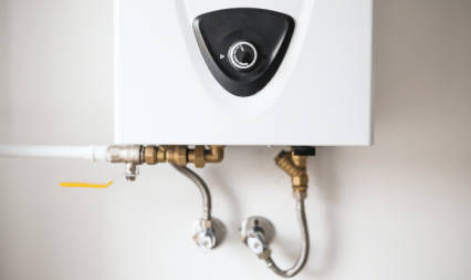 Close-up of a white tankless water heater with control dial, attached to wall-mounted plumbing fittings.