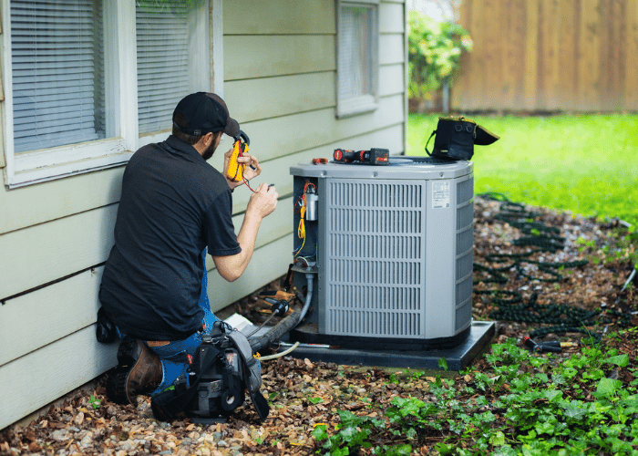 HVAC technician performing maintenance on an outdoor air conditioning unit beside a house, using tools and diagnostic equipment to check system functionality.