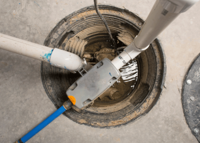 Top-down view of a sump pit with active plumbing connections and a sump pump installed, showing water accumulation and drainage components in a basement or utility area.