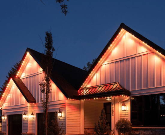 Modern home with white siding and black rooflines illuminated by Gemstone Lights in a warm orange glow, highlighting roof peaks and architectural details at dusk.