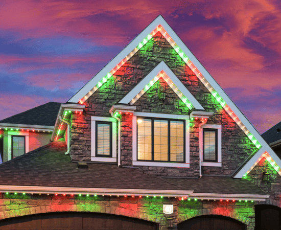 Stone exterior home with Gemstone Lights in red and green holiday colors outlining the roof peaks and eaves at sunset.