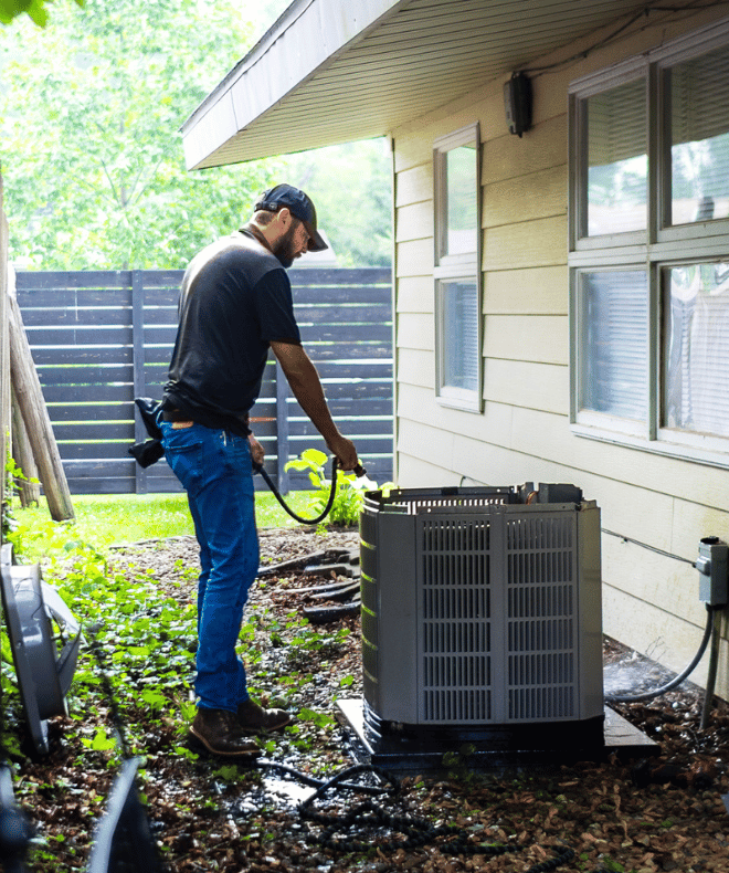 An HVAC technician is spraying down a residential AC condenser unit during routine maintenance on the side of a house.