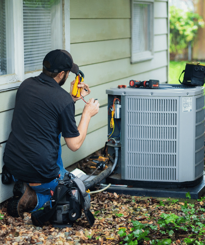 A technician kneels beside a residential home, testing an outdoor air conditioning unit with a clamp meter. The unit's access panel is open, revealing wiring and components, while tools and a vacuum pump sit nearby. The technician is conducting HVAC repair work in a shaded yard area.