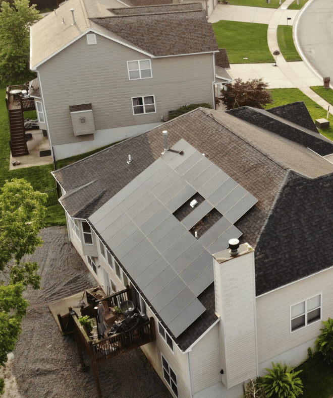 Aerial view of a suburban home with solar panels installed across the main roof slope. The panels are arranged around existing roof vents and chimneys, maximizing available space. Surrounding homes and a curved residential street are visible in the background.