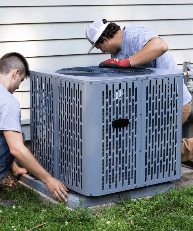 Two technicians install or inspect an outdoor air conditioning unit next to a house. One adjusts the concrete base while the other examines the top of the unit, ensuring it's level and properly positioned. The scene captures a residential HVAC setup in progress.