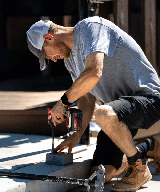Electrician installing an outdoor electrical box with a Milwaukee power drill, wearing a gray C-U Trade Services shirt, black shorts, and work boots/