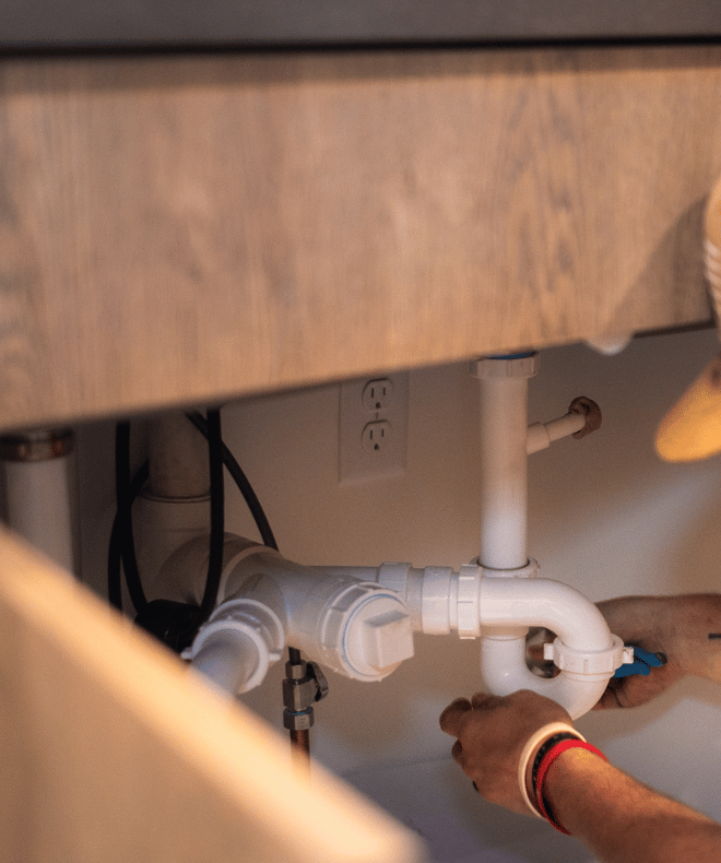 Plumber tightening a PVC drain trap under a wooden vanity cabinet in a residential bathroom.