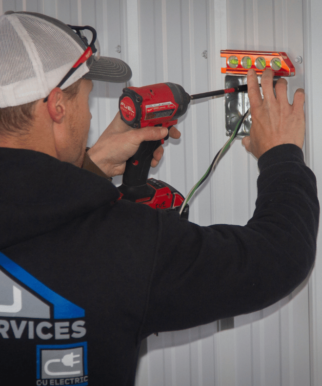 A technician installs a wall-mounted electrical box using a power drill while checking alignment with a level. He wears a C-U Trade Services hoodie featuring the C-U Electric logo, working on a metal-paneled wall during a residential or light commercial electrical project.