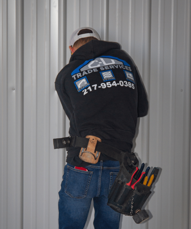 A C-U Trade Services technician installs wiring or equipment on a metal-paneled wall. The back of his hoodie clearly displays the company’s branding and phone number (217-954-0385), with icons for plumbing, HVAC, and electrical services. A loaded tool belt and holstered hammer emphasize hands-on expertise.