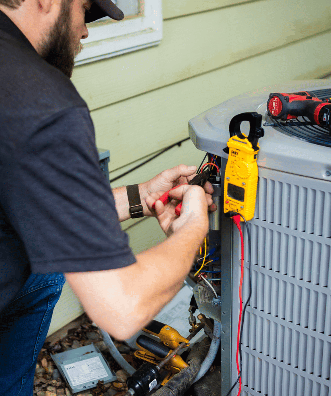 A technician uses pliers to work on electrical wiring inside an outdoor air conditioning unit. A multimeter and tools are positioned nearby, indicating active HVAC troubleshooting or repair on a residential property.