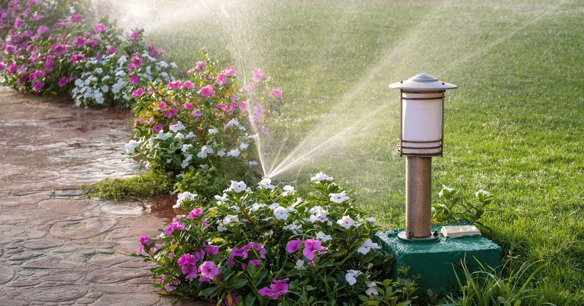 Sprinkler system watering a landscaped garden with blooming pink and white flowers along a stone path, next to a metal outdoor light fixture on a green lawn.