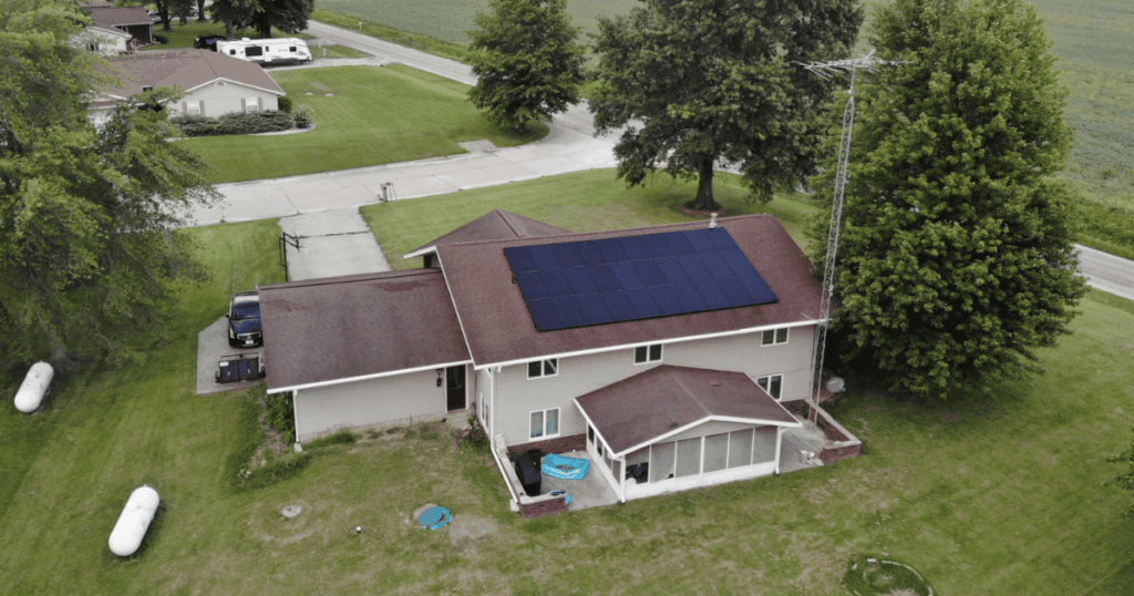 A two-story suburban home with dark rooftop solar panels installed on one side of the roof. The house has a red-brown shingle roof, a screened-in porch addition, a driveway with a parked vehicle, and large trees surrounding the property.