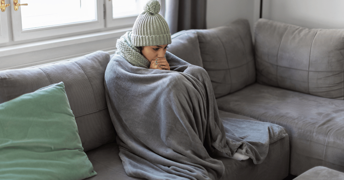 **Alt text:** A person bundled in a gray blanket and winter hat sits on a couch looking cold, showing what it feels like when the heater stops working at home.