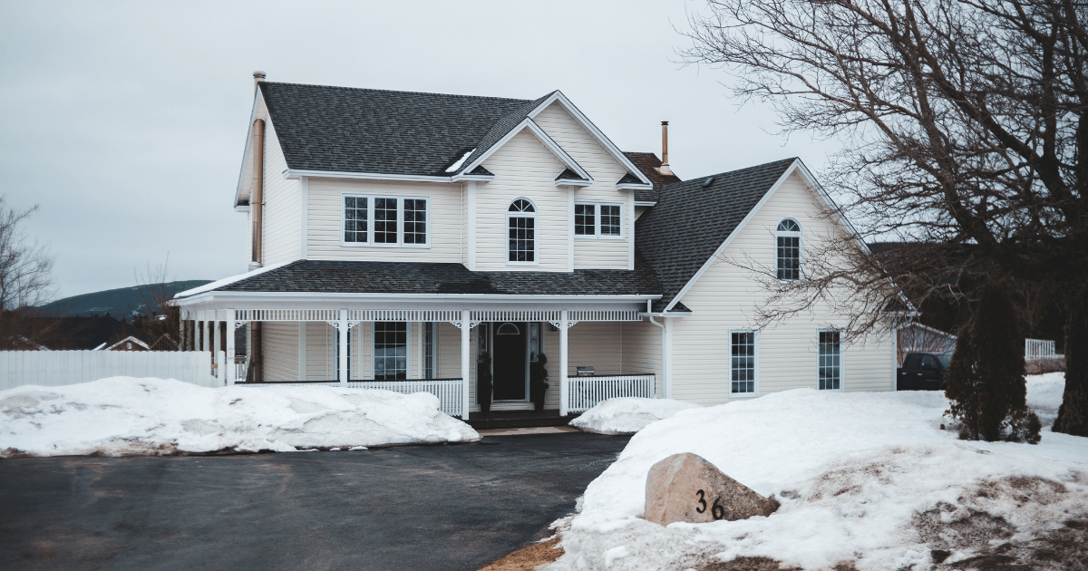 Two-story beige house with a dark gray roof surrounded by patches of melting snow, a bare tree in the yard, and overcast winter skies in a suburban neighborhood.